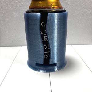 May include: A blue can cooler with a can inside. The cooler has a cylindrical shape with a slot cut out to reveal the can's label. The can is gold and black. The cooler has the text "e N R DI 23.2".