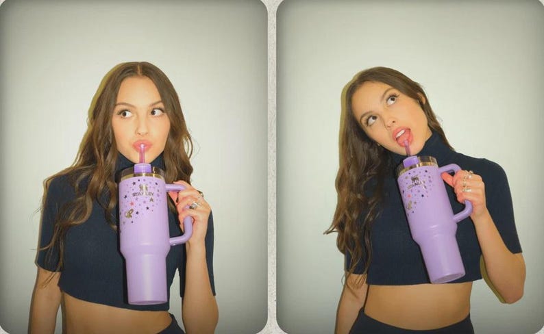 May include: A light purple insulated tumbler with a handle, a straw, and a decorative design. The tumbler is held by a person wearing a dark blue crop top. The tumbler has the text "Stanley" on it.