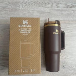May include: A brown Stanley Flowstate tumbler with a handle and straw, next to its cardboard box. The box features the Stanley logo and product details, including "40 oz". The tumbler is designed to keep drinks cold for 2 days and hot for 7 hours.