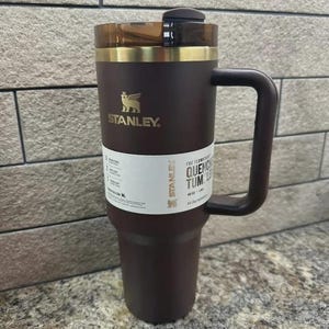 May include: A dark brown Stanley Quencher tumbler with a handle and a clear lid. The tumbler has a gold band around the top and the Stanley logo in gold. A white label with text is wrapped around the tumbler.
