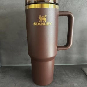 May include: A brown Stanley travel mug with a handle, a clear lid, and a gold rim. The Stanley logo is displayed in gold. The mug has a straw and is designed to keep beverages hot or cold.