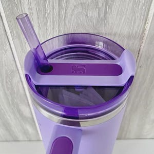 May include: A purple insulated tumbler with a handle and a clear lid. The lid has a sliding closure and a purple straw. The tumbler has a silver band near the top and a logo on the lid.
