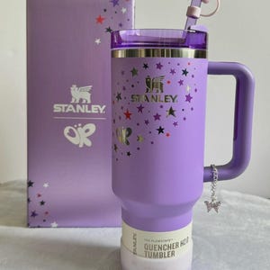 May include: A lavender Stanley Quencher H2.0 Tumbler with a handle, straw, and star-shaped topper. The tumbler is decorated with stars and butterflies. A matching box with the Stanley logo and butterfly graphic is behind the tumbler.