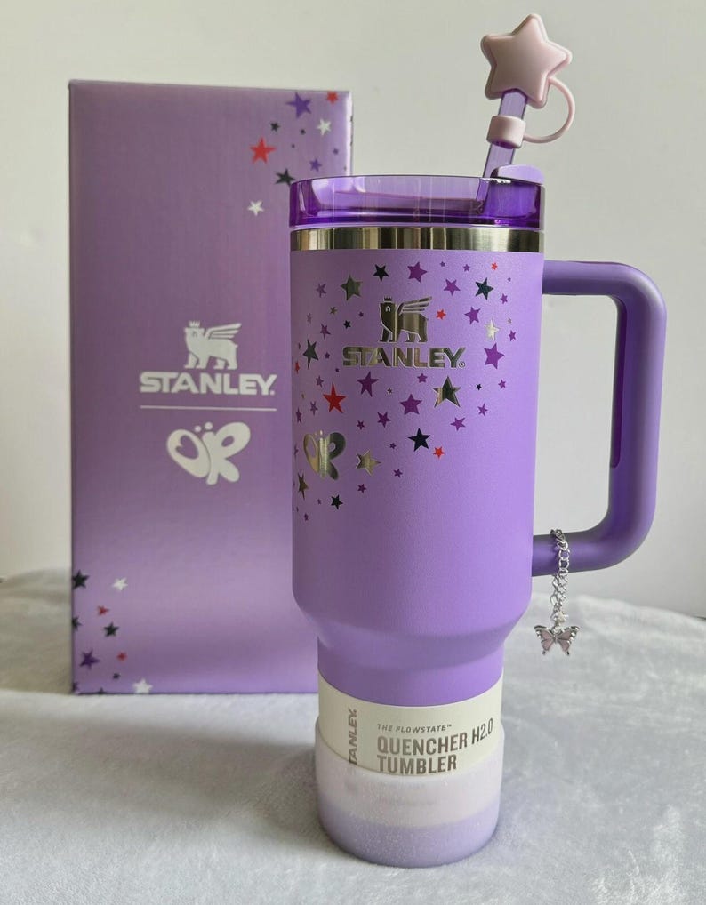 May include: A lavender Stanley Quencher H2.0 Tumbler with a handle, straw, and star-shaped topper. The tumbler is decorated with stars and butterflies. A matching box with the Stanley logo and butterfly graphic is behind the tumbler.