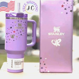 May include: A lavender Stanley Quencher H2.0 tumbler with a handle, straw, and lid. The tumbler is decorated with gold and red stars and a butterfly graphic. The box next to it is lavender and has the Stanley logo and a butterfly graphic.