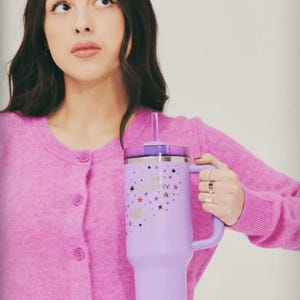 May include: A lavender insulated tumbler with a handle and a straw. The tumbler features a design of stars and the text "Good 4 U Honey". The tumbler is held by a person wearing a pink cardigan.