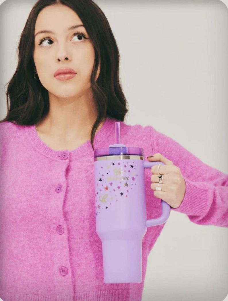 May include: A lavender insulated tumbler with a handle and a straw. The tumbler features a design of stars and the text "Good 4 U Honey". The tumbler is held by a person wearing a pink cardigan.