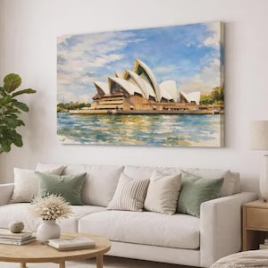 May include: A canvas print of the Sydney Opera House hangs above a white sofa with green and beige pillows. The artwork depicts the iconic building with a blue sky and water. A round wooden coffee table and a potted plant are in front of the sofa.