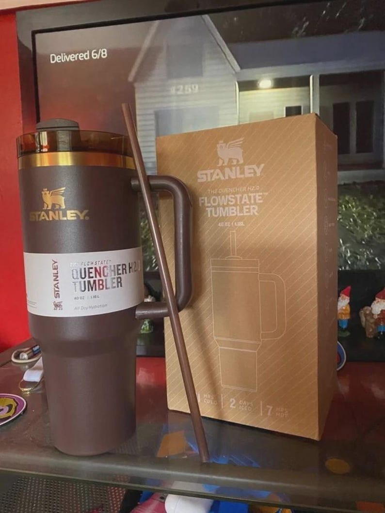 May include: A brown Stanley Flowstate Quencher H2.0 Tumbler with a handle and straw, next to its cardboard box. The box features the Stanley logo and product details. The tumbler holds 40 oz.