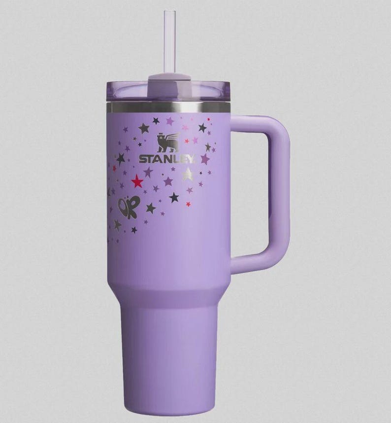 May include: A lavender Stanley tumbler with a handle and a straw. The tumbler is decorated with a pattern of stars and butterflies. The Stanley logo is visible on the side.