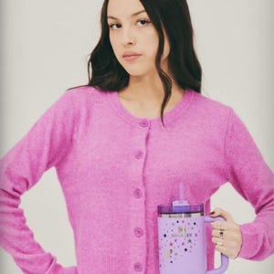 May include: A person wearing a pink cardigan holding a large, lavender Stanley tumbler. The tumbler features a handle, a straw, and a design of stars and the Stanley logo. The cardigan has a button-up front.