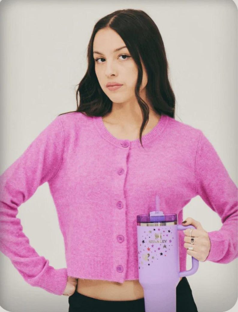May include: A person wearing a pink cardigan holding a large, lavender Stanley tumbler. The tumbler features a handle, a straw, and a design of stars and the Stanley logo. The cardigan has a button-up front.