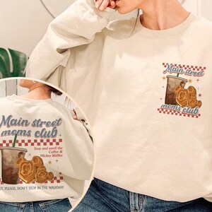 Pu&ograve; includere: Felpa color crema con la scritta "Main street moms club" in blu e rosso. Il design include una tazza di caff&egrave; e waffle di Mickey Mouse. Il retro della felpa ripete il design con la scritta "Stop and smell the Coffee & Mickey Waffles."