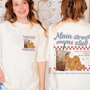 Pu&ograve; includere: T-shirt color crema con grafica di caff&egrave; e waffle di Mickey Mouse. La parte anteriore della maglietta dice "Main Street Moms Club". La parte posteriore della maglietta ha la stessa grafica e dice "Stop and smell the Coffee & Mickey Waffles."