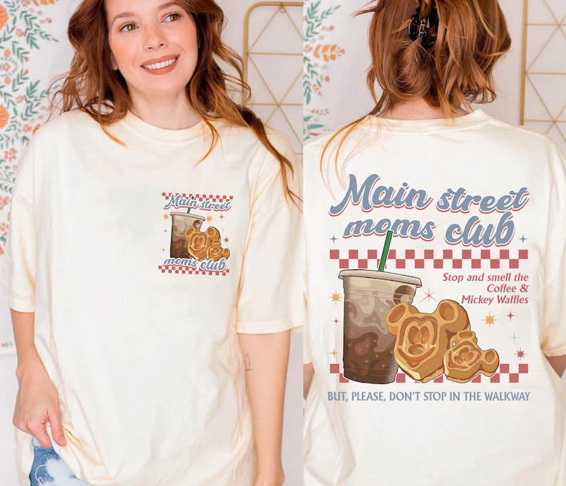 Pu&ograve; includere: T-shirt color crema con grafica di caff&egrave; e waffle di Mickey Mouse. La parte anteriore della maglietta dice "Main Street Moms Club". La parte posteriore della maglietta ha la stessa grafica e dice "Stop and smell the Coffee & Mickey Waffles."