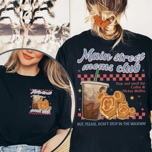 Pu&ograve; includere: T-shirt nero con la scritta "Main Street Moms Club" e grafica. Il design include una tazza di caff&egrave;, waffle di Mickey Mouse e la frase "Stop and smell the Coffee & Mickey Waffles."