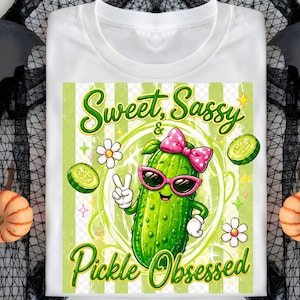 Könnte beinhalten: Weißes T-Shirt mit einer Cartoon-Gurke, die eine rosa Sonnenbrille und eine Schleife trägt und ein Friedenszeichen gibt. Das Shirt trägt die Worte "Sweet, Sassy & Pickle Obsessed" in Grün und Gelb.