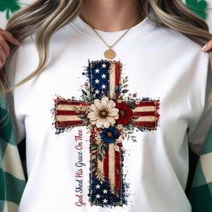 Può includere: Felpa bianca con un design a croce con un motivo a bandiera americana. La croce è adornata da accenti floreali rossi, bianchi e blu, e il testo "God Shed His Grace On Thee" è stampato verticalmente sul lato sinistro.