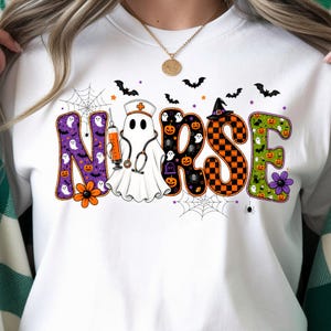 May include: A white crewneck sweatshirt featuring the word NURSE in large, colorful letters. The letter U is replaced by a ghost wearing a nurse cap and stethoscope, holding a syringe. The design includes pumpkins, bats, spiders, and webs.