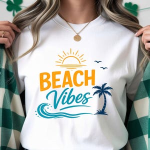 Op de afbeelding: Wit T-shirt met ronde hals en de tekst "BEACH Vibes" in oranje en blauwgroen. Het ontwerp bevat een zon, golven, een palmboom en vogels. Een casual, comfortabele stijl.
