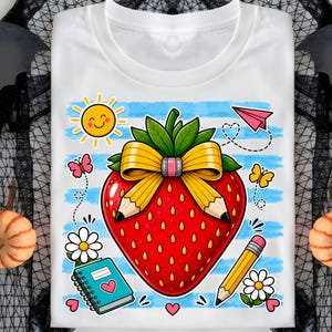 Op de afbeelding: Wit T-shirt met een ontwerp van een rode aardbei met een gele strik en potloden. Het ontwerp bevat een lachende zon, vlinders, een papieren vliegtuigje, een notitieboekje en madeliefjes. Blauw-wit gestreepte achtergrond.