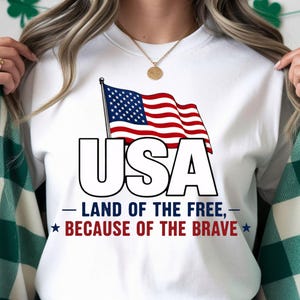 Op de afbeelding: Wit T-shirt met een afbeelding van de Amerikaanse vlag en het woord "USA". Onder de vlag staat de tekst "Land of the Free, Because of the Brave" in rood en blauw. Een patriottisch ontwerp.