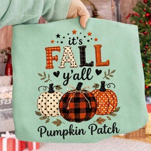 May include: A light mint green sweatshirt featuring a graphic with the text "it's FALL y'all Pumpkin Patch". The design includes three pumpkins with different patterns: a white polka dot pumpkin, a red and black buffalo plaid pumpkin, and an orange polka dot pumpkin, accented with small stars, hearts, and leaves.