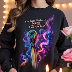 May include: Black sweatshirt featuring the text "Your Best Teacher Is Your Last Mistake" and a colourful graphic of a burnt match. The match head is a vibrant mix of colours, with swirling, colourful smoke-like designs around it.