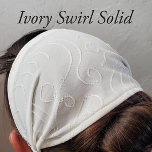 Puede incluir: Diadema de color marfil con un diseño de remolino. La tela es de color marfil liso con remolinos bordados. El texto "Ivory Swirl Solid" se muestra en la imagen. La diadema está diseñada para ser usada en la cabeza.