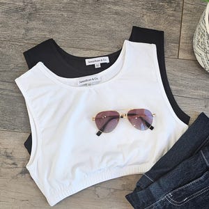 May include: A white and black crop top with the text "Jamie Rose & Co" and a pair of aviator sunglasses with a gold frame and pink lenses. Dark denim jeans are also in the image.