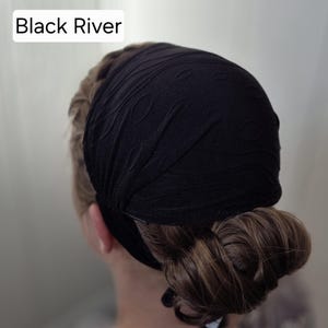 Può includere: Una fascia nera con un motivo testurizzato a vortice sul tessuto. Il testo Black River è visibile nell'angolo in alto a sinistra. La fascia è indossata intorno alla testa, copre la corona ed è fissata sulla parte posteriore della testa.