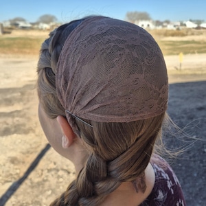 May include: A brown lace head covering, secured with a metal pin, is worn over the hair. The hair is styled in a braid. The head covering has a floral lace pattern. The background is blurred, with a clear blue sky.