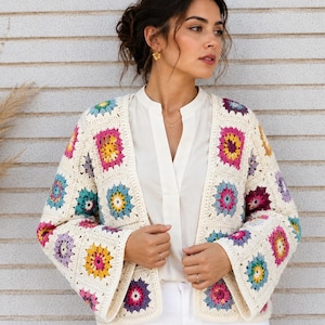 Op de afbeelding: Een gehaakt vest opgebouwd uit gebroken witte granny squares met kleurrijke bloemcentra in tinten geel, roze, paars en groenblauw. Het kledingstuk heeft een open voorkant, lange mouwen en een vierkant patroon over het hele lichaam.