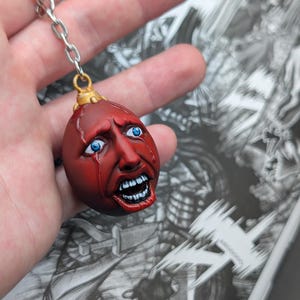 May include: A red, egg-shaped keychain pendant with a detailed, anguished face, blue eyes, and open mouth. The pendant is attached to a silver chain. The background features black and white manga illustrations.