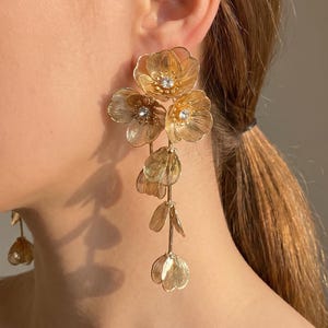 May include: A long gold-toned earring featuring a cluster of translucent, layered flower petals arranged in a cascading vertical design. Each flower centre is accented with a small, clear, sparkling crystal, and the petals have a delicate, veined texture.