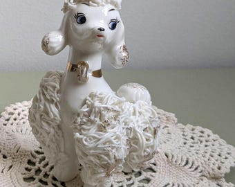 May include: A white ceramic poodle figurine featuring textured fur, a gold-tone collar, and a small pink rose accent on its head. The poodle has painted blue eyes, black eyelashes, and gold-tone detailing on its ears and tail.