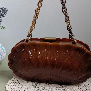 May include: A brown marbled acrylic handbag with a shell-like fluted design and a rectangular metal clasp. The bag features a long, decorative gold-tone chain handle with interlocking circular links that transition to a darker brown tone.