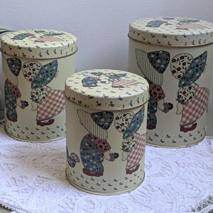 May include: A set of three cylindrical metal canisters in varying heights, featuring a cream-coloured background with a repeating pattern of figures wearing bonnets and checkered dresses, complemented by a white ceramic vase with floral accents.