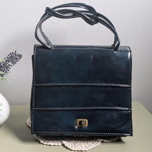 May include: A dark navy blue patent leather handbag with a glossy finish, featuring a structured rectangular shape, two top handles, and a gold-toned metal turn-lock closure on the front flap. The bag has horizontal stitched detailing across the front.