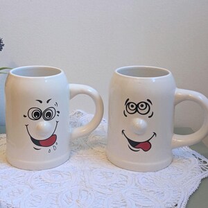Peut inclure: Deux tasses en céramique blanche dotées de nez en relief en trois dimensions et d'expressions faciales imprimées en noir et blanc avec des langues rouges tirées. Chaque tasse possède une anse latérale robuste et un corps cylindrique lisse.