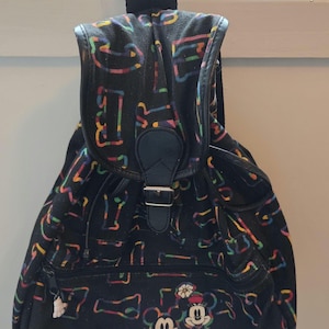 May include: A black backpack featuring a colorful, repeating abstract pattern and a front graphic of Mickey and Minnie Mouse. The bag includes a flap closure with a black buckle, a zippered front pocket with a small white glove zipper pull, and straps.