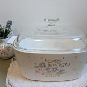 Puede incluir: Fuente blanca para horno con tapa de cristal transparente. El plato presenta un diseño de cesta floral con flores rosas y grises. A la izquierda hay un pequeño jarrón con lavanda y un diseño floral.