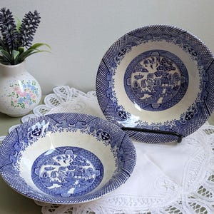 May include: Two round ceramic bowls featuring a blue and white willow pattern with a detailed landscape scene in the center and a geometric border. The bowls have a cream-colored base and a glossy finish, showcasing a classic transferware design.