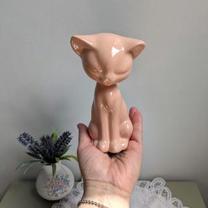 May include: A peach-coloured ceramic cat figurine with a smooth, glossy finish. The cat is in a seated position with its eyes closed and ears perked up. The figurine is held in a hand, showcasing its size and detail. A decorative vase with flowers is in the background.