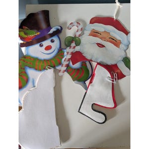 May include: A decorative cutout featuring a smiling snowman wearing a black top hat with a small bird perched on the brim, alongside a cheerful Santa Claus holding a red and white striped candy cane. Both figures have green striped scarves.
