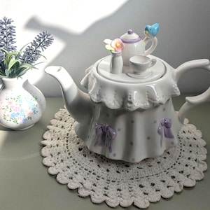 May include: A white ceramic teapot with a ruffled skirt design, decorated with lavender bows and polka dots. A miniature teapot, teacup, and vase with flowers sit on the lid. A small blue bird decorates the teapot. A vase of lavender is on the left.