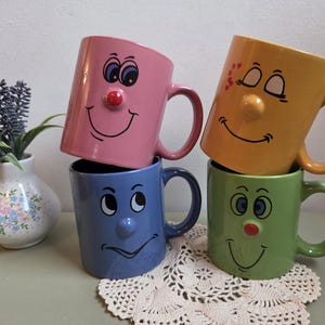 Peut inclure: Quatre tasses en céramique rose, jaune, bleue et verte, chacune dotée d'un nez en relief et d'un visage imprimé avec des yeux et un sourire. Les tasses rose et verte ont des nez rouges, tandis que les jaunes et bleues ont des nez assortis.