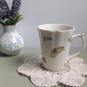May include: A cream-colored ceramic mug with a scalloped rim and gold-tone trim, featuring a delicate butterfly and small floral pattern. The mug has a single handle and sits on a round, off-white crocheted doily next to a small floral vase.