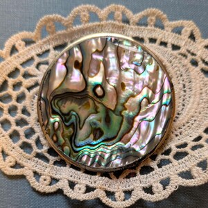 May include: A round, iridescent abalone shell compact with a silver-colored metal rim, resting on a cream-colored, openwork lace doily. The shell displays a mix of colors, including green, blue, and pink. The doily is placed on a blue surface.