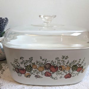 May include: A white casserole dish with a clear glass lid. The dish has a colourful vegetable and herb design around the base. A small vase with lavender and a decorative vase are in the background.